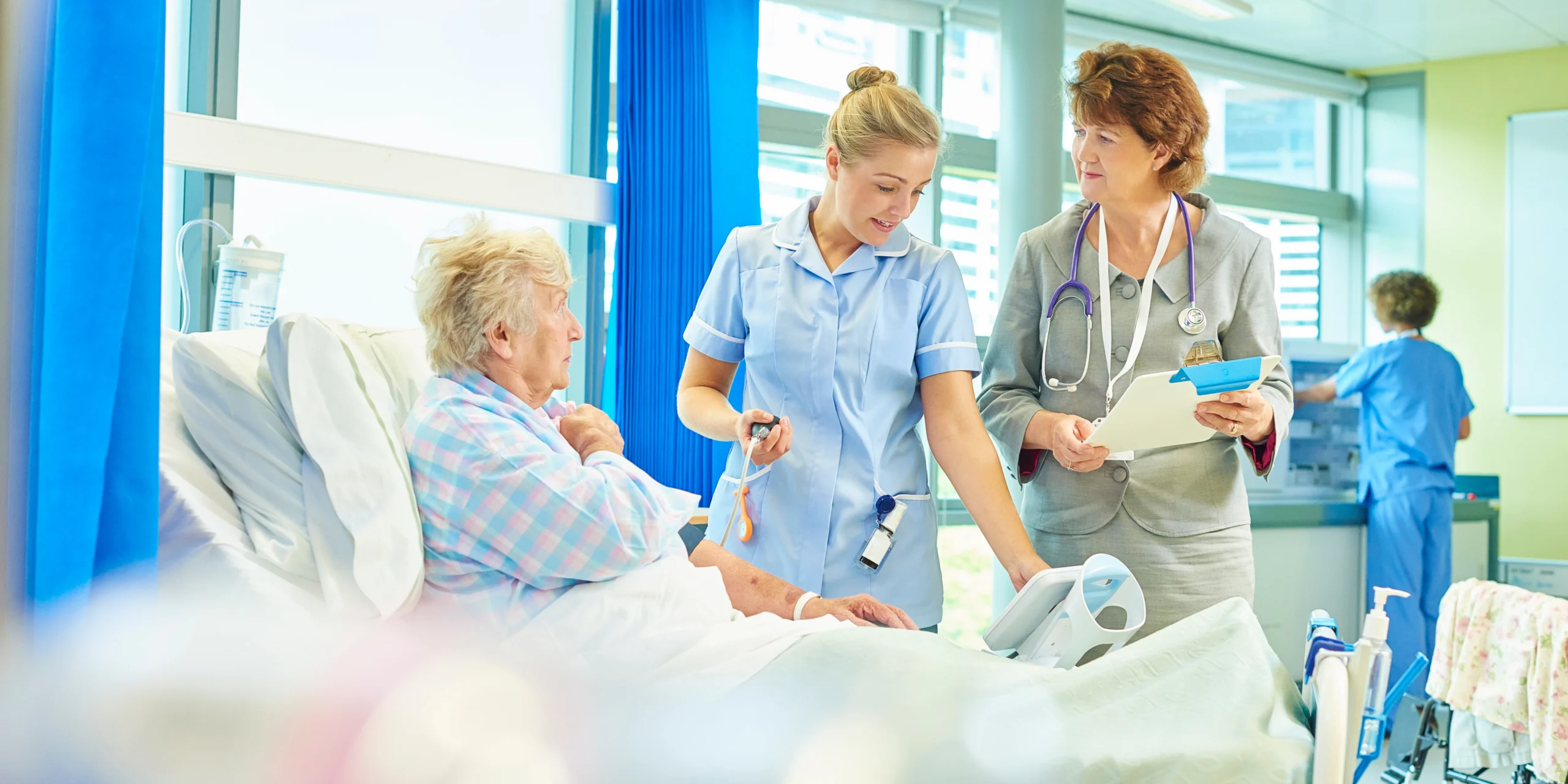 Elderly female patient in hospital bed being attended to by a nurse in blue uniform and a doctor in grey suit holding a clipboard in a hospital ward